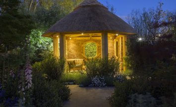 Garden Lighting. Wooden chair in thatched summerhouse lit at night. Bespoke Summerhouse hand-crafted from Oak